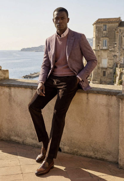A man wearing a pink and brown checkered blazer, a pink knit sweater, dark brown trousers, and brown leather loafers, leaning against a stone wall with a coastal town and sea in the background.