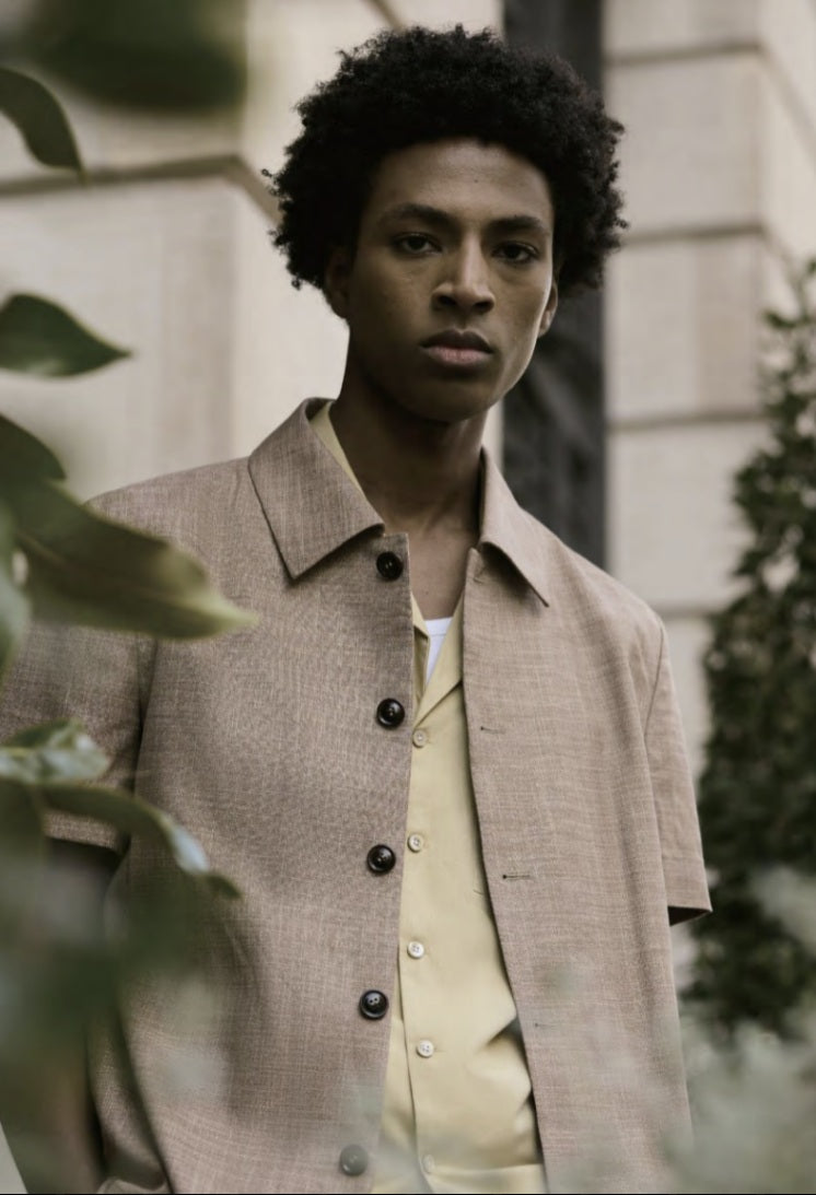 A man wearing a short-sleeved, button-up shirt in a sand color, with a collar, made of a blend of bamboo, spandex, silk, and wool fabrics.