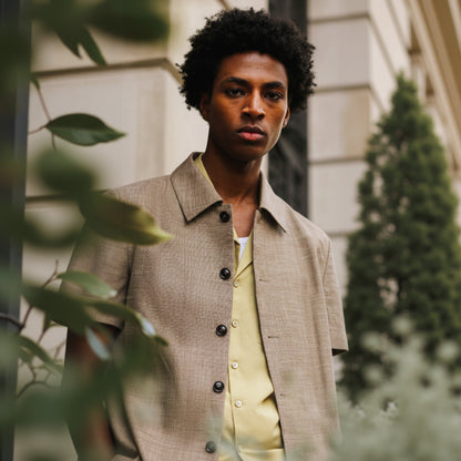 A man wearing a short-sleeved, button-up shirt in a sand color, with a collar, made of a blend of bamboo, spandex, silk, and wool fabrics.