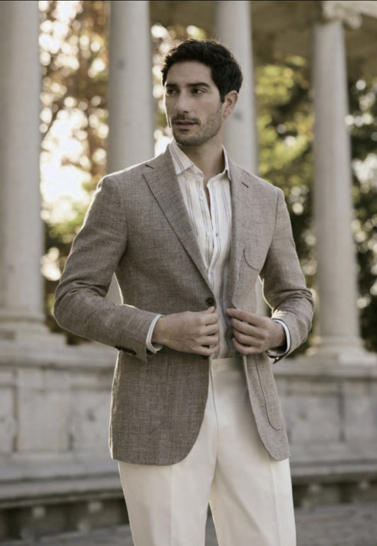 A man wearing a taupe linen-cotton jacket with dark brown buttons and white trousers standing in a columned hallway.