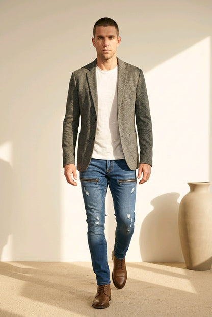 A man wearing a textured gray blazer, white t-shirt, distressed blue jeans, and brown leather shoes walking in a minimalist beige room with a large ceramic vase.