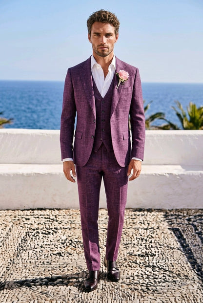 A man wearing a textured purple three-piece suit with a white shirt and a pink boutonniere, standing outdoors on a cobblestone surface with the ocean and white wall in the background.