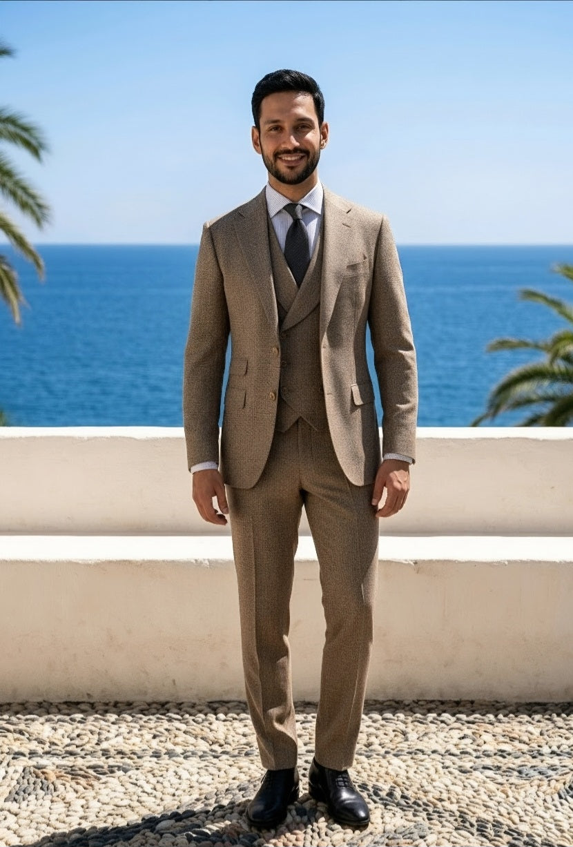 A man wearing a three-piece beige tweed suit with a white shirt and dark tie, standing outdoors in front of a white wall and the blue ocean.