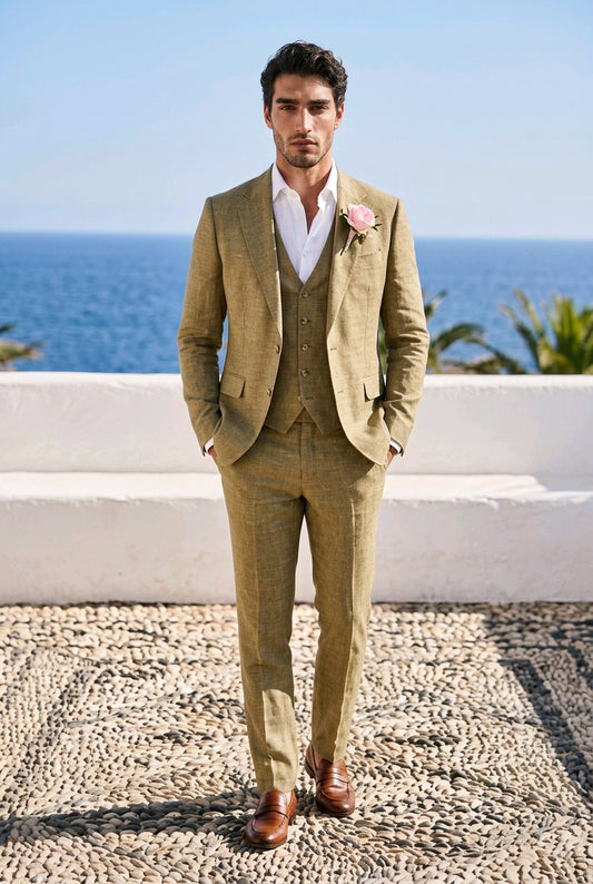 A man wearing a three-piece olive green tweed suit with a white shirt and a pink boutonniere, standing outdoors in front of a white wall and the blue ocean.