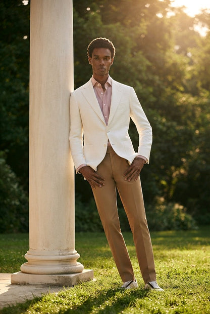 A man wearing a white blazer, tan trousers, and a pink shirt stands outdoors next to a large white column in a sunlit garden setting.