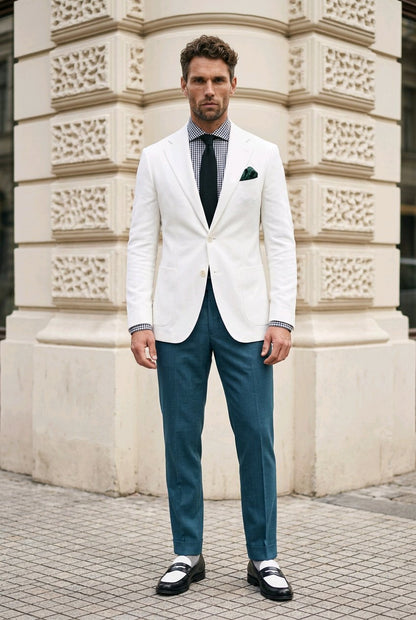 A man wearing a white blazer, teal trousers, a dark tie, and a patterned dress shirt stands in front of a beige stone building.