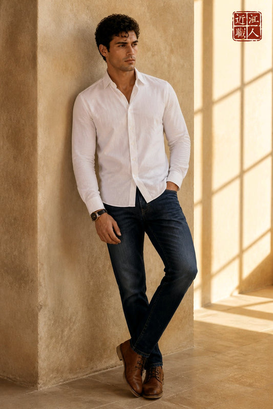 A man wearing a white long-sleeve button-down shirt, dark blue jeans, and brown leather shoes leans against a textured beige wall in a sunlit room with window shadows.