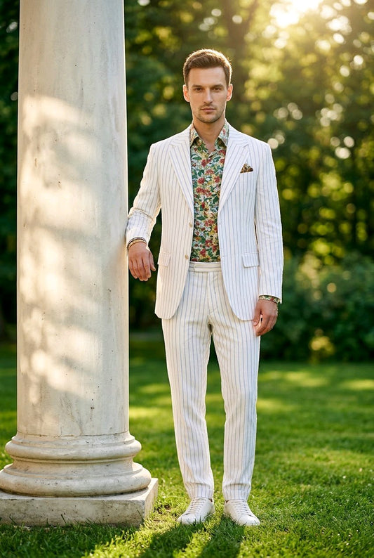 A man wearing a white pinstripe suit with a colorful floral shirt and white shoes, standing outdoors next to a large white column on a grassy lawn with trees in the background.