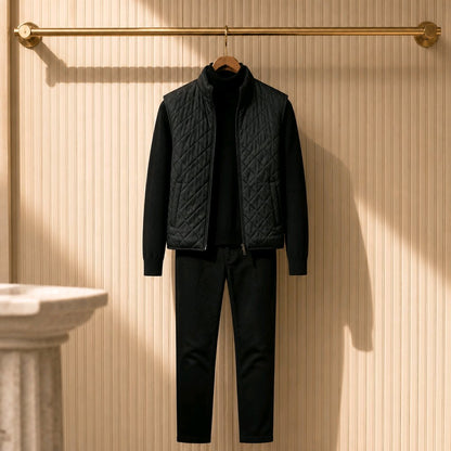 A men's black quilted vest layered over a black zip-up sweater and matching black trousers, hanging on a wooden hanger from a gold wall-mounted rod against a beige ribbed wall.