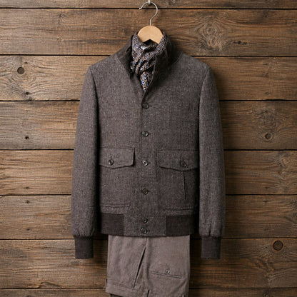 A men's dark brown textured wool-blend cardigan sweater with a high collar, button front, and flap pockets, styled with a patterned scarf and matching brown trousers against a rustic wooden plank background.