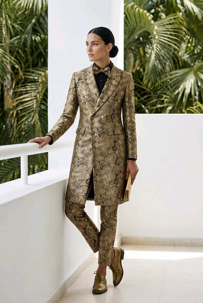 A model stands on a white balcony wearing a matching snakeskin-patterned blazer and trousers suit in shades of brown and beige, paired with a black shirt and gold shoes.