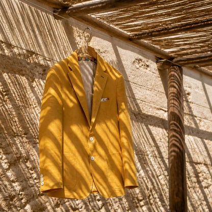 A mustard yellow linen blazer jacket hanging on a wooden hanger against a rustic sunlit wall with shadows from a thatched roof.
