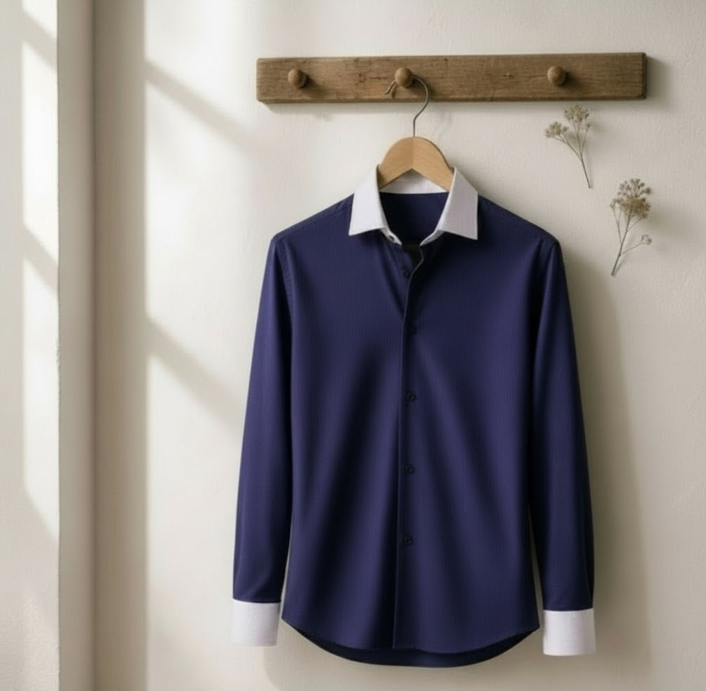 A navy blue long-sleeve button-up shirt with a contrasting white collar and white cuffs, hanging on a wooden hanger from a rustic wooden wall hook against a warm off-white wall.