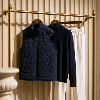 A navy quilted zip-front vest hanging on a brass rack, with a matching navy knit turtleneck sweater and white pants displayed beside it against a beige slatted wall.