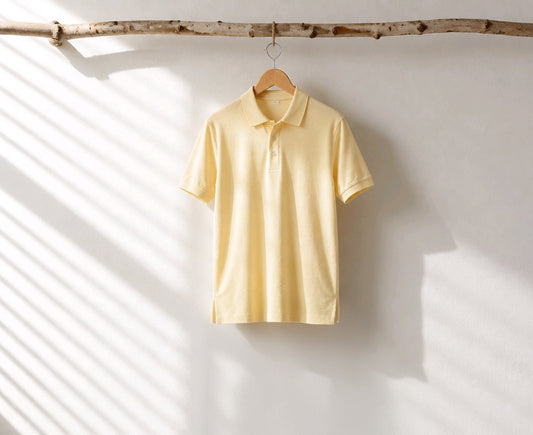 A pale yellow short-sleeve polo shirt hanging on a wooden hanger from a rustic branch, set against a white wall with soft diagonal shadows.