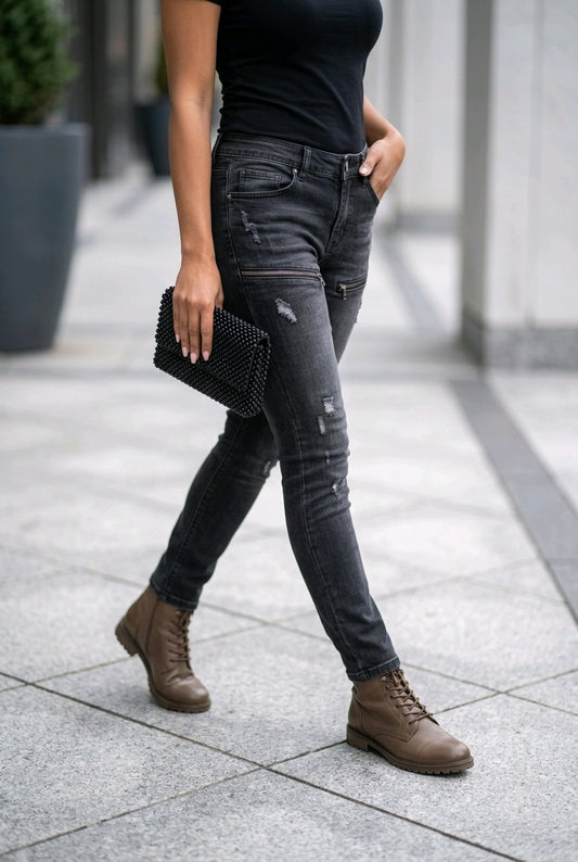 A person walking outdoors wearing dark gray distressed skinny jeans, a black top, brown lace-up ankle boots, and holding a black textured clutch.
