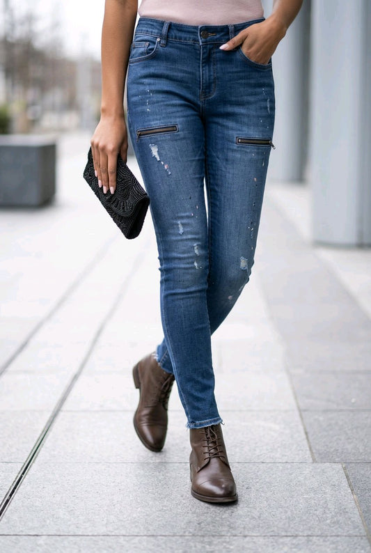 A person walking outdoors wearing medium-wash blue skinny jeans with distressed details and zippered pockets on the thighs, paired with brown ankle boots and holding a black clutch.