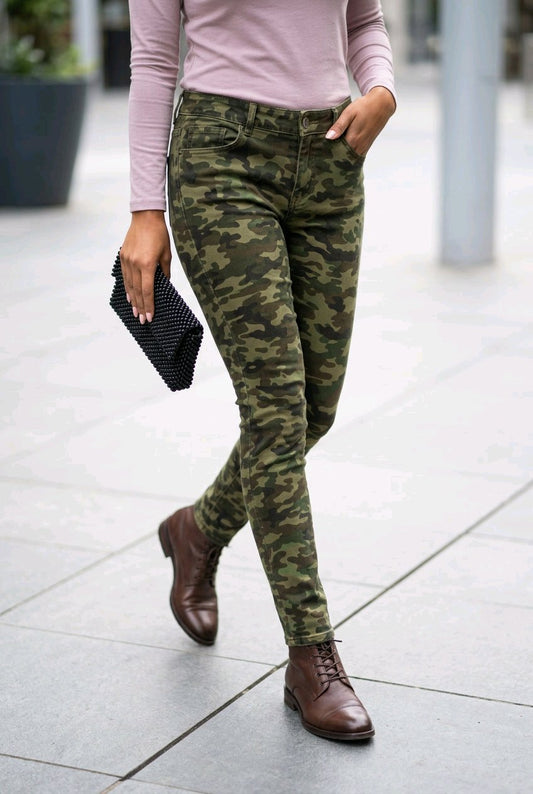 A person walking outdoors wearing slim-fit camouflage pants, a light pink long-sleeve top, and brown leather ankle boots, while holding a black patterned clutch.