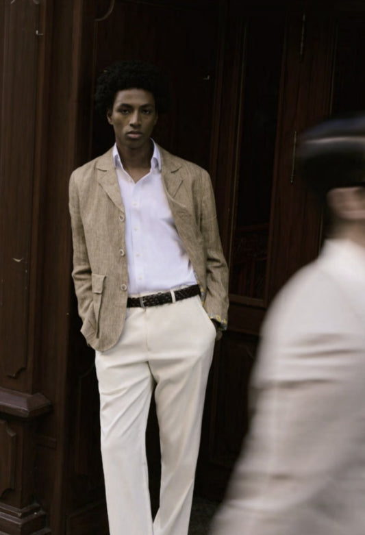 A person wearing a beige jacket over a white shirt paired with white trousers standing against a wooden background.