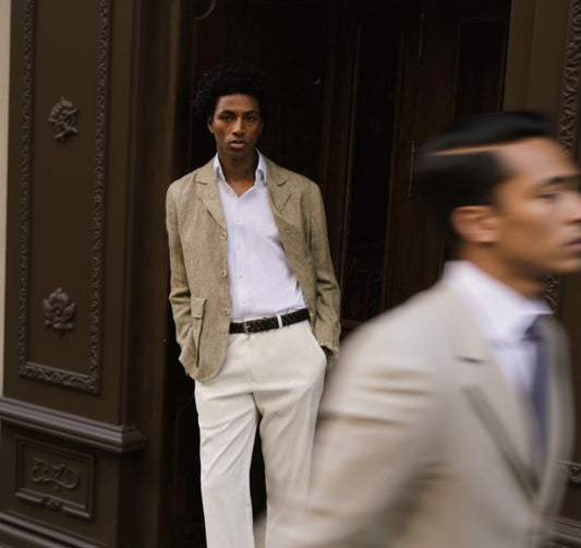 A person wearing a beige jacket over a white shirt paired with white trousers standing against a wooden background.