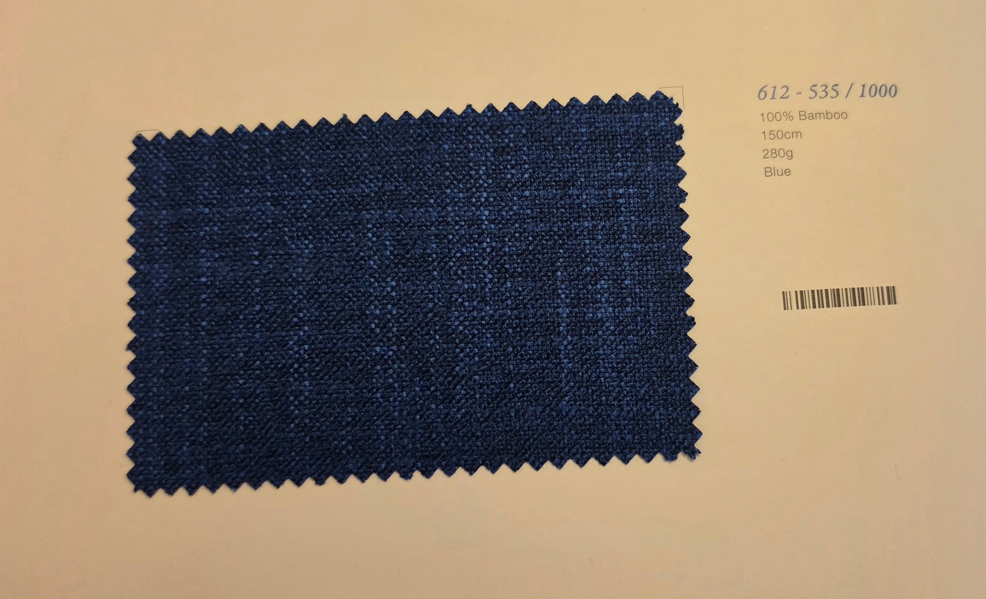 A rectangular fabric swatch of dark blue woven material with zigzag-cut edges, placed on a light beige background with printed text and a barcode to the right.