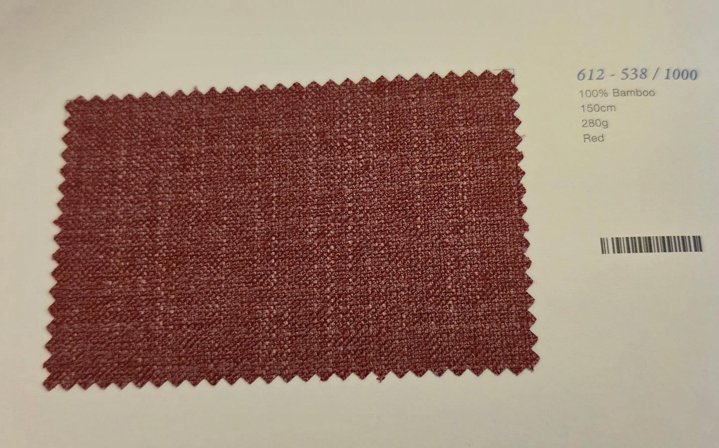A rectangular woven fabric swatch in a muted brick red color with zigzag-cut edges, displayed on a light beige background with printed text and a barcode on the right.