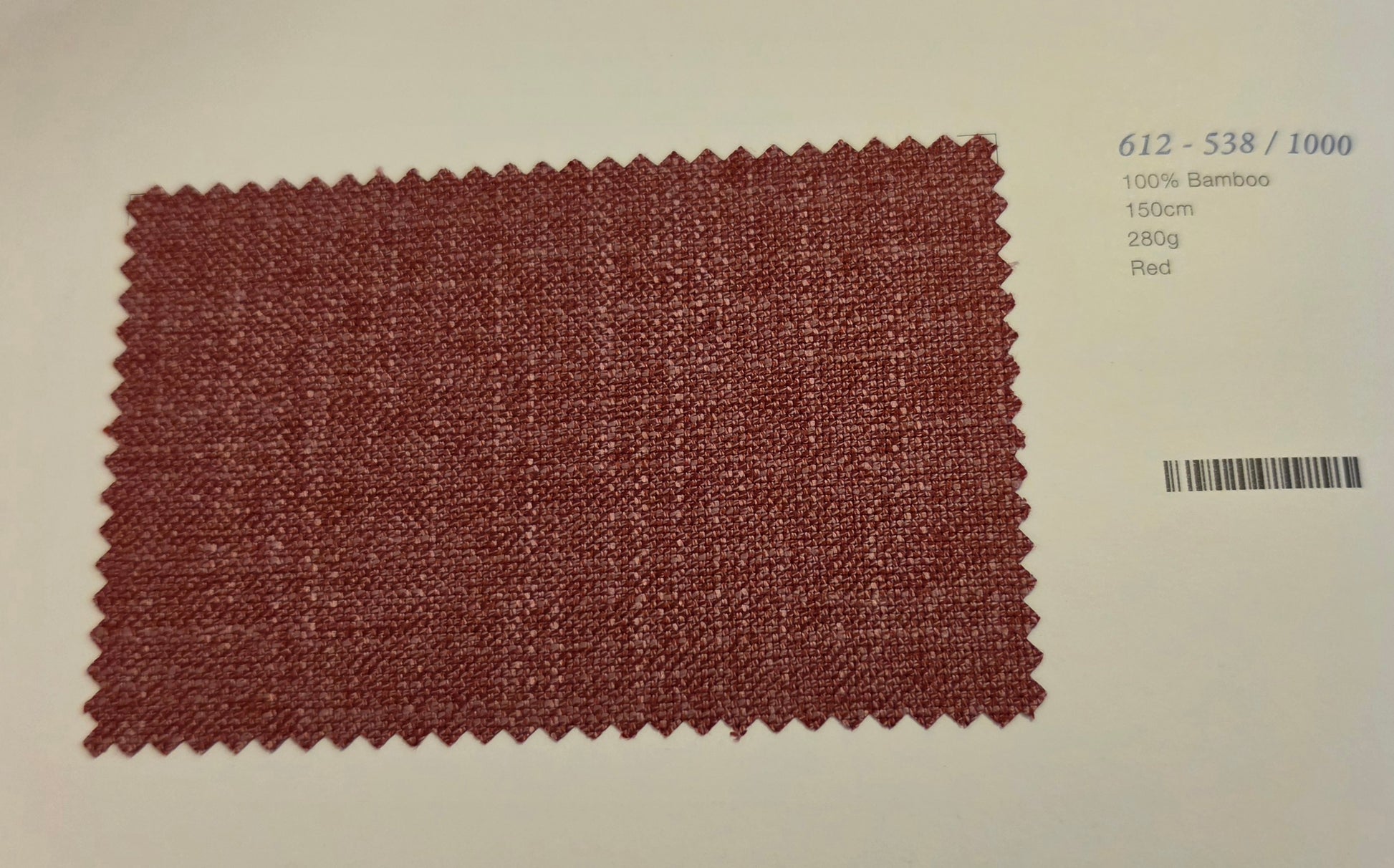 A rectangular woven fabric swatch in a muted brick red color with zigzag-cut edges, displayed on a light beige background with printed text and a barcode on the right.