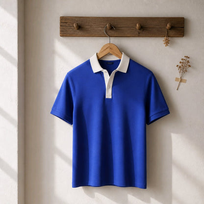 A royal blue short-sleeve polo shirt with a white collar and white placket, hanging on a wooden hanger from a rustic wooden wall hook. The shirt is displayed against a warm, off-white textured wall with soft diagonal shadows.