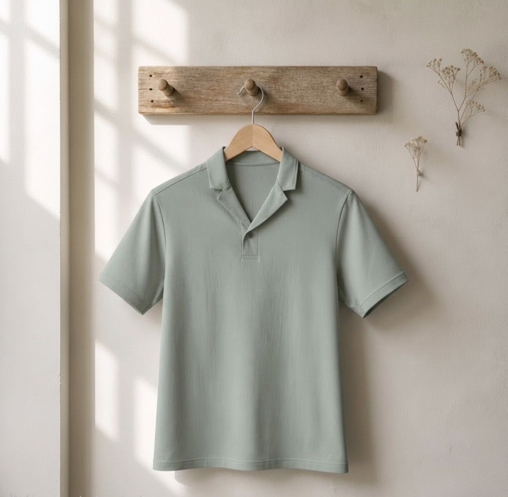 A sage green short-sleeve polo shirt hanging on a wooden hanger from a rustic wooden peg rail against a warm beige wall with soft window light shadows.