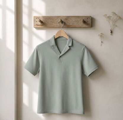 A sage green short-sleeve polo shirt hanging on a wooden hanger from a rustic wooden peg rail against a warm beige wall with soft window light shadows.