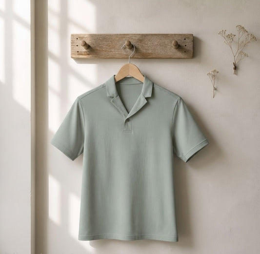 A sage green short-sleeve polo shirt hanging on a wooden hanger from a rustic wooden peg rail against a warm beige wall with soft window light shadows.