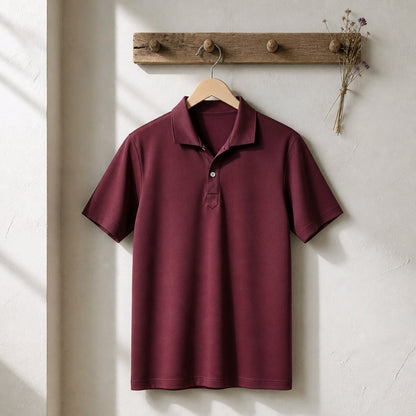 A short-sleeve polo shirt in deep burgundy is hanging on a wooden hanger from a rustic wooden peg rack against a textured off-white wall.