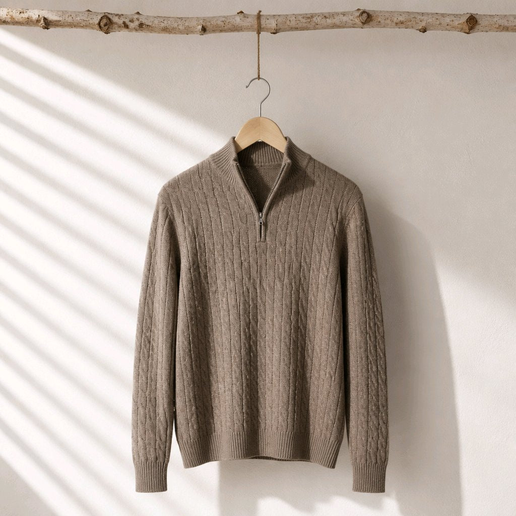 A taupe cable-knit half-zip sweater hanging on a wooden hanger from a rustic branch, set against a white wall with soft diagonal shadow patterns.