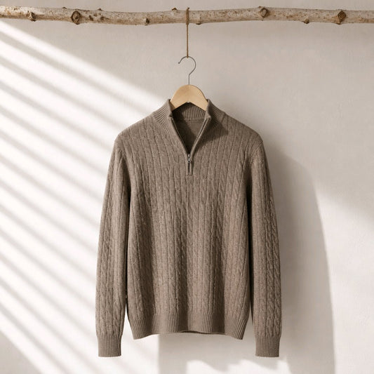 A taupe cable-knit half-zip sweater hanging on a wooden hanger from a rustic branch, set against a white wall with soft diagonal shadow patterns.