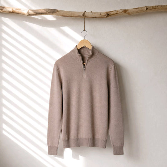 A taupe knit quarter-zip sweater hanging on a wooden hanger from a rustic branch, set against a textured off-white wall with soft diagonal window shadow patterns.