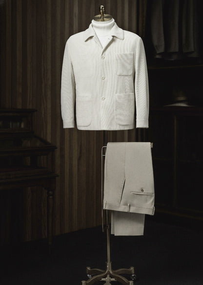A white cord casual jacket and beige trousers displayed on a mannequin, with the jacket featuring a high shirt collar, three-button fastening, and round patch breast pocket, and the trousers having a plain waistband and front pockets.