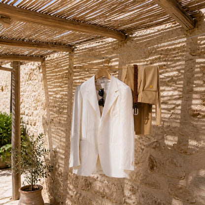A white linen blazer and tan trousers hang on wooden hangers on a rustic stone wall under a bamboo roof, with a brown leather belt and sunglasses as accessories.