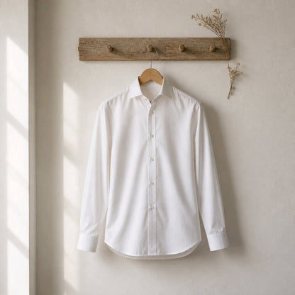 A white long-sleeve button-up dress shirt hanging on a wooden hanger from a rustic wooden wall hook, set against a warm beige textured wall with soft window light casting shadows.