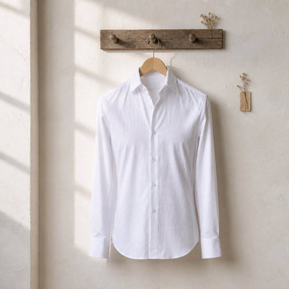 A white long-sleeve button-up shirt hangs on a wooden hanger from a rustic wooden wall rack, set against a textured beige wall with soft window shadow light.