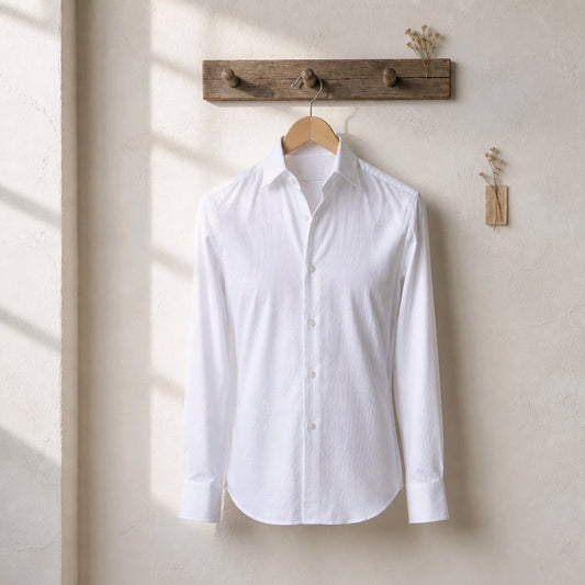 A white long-sleeve button-up shirt hangs on a wooden hanger from a rustic wooden wall rack, set against a textured beige wall with soft window shadow light.