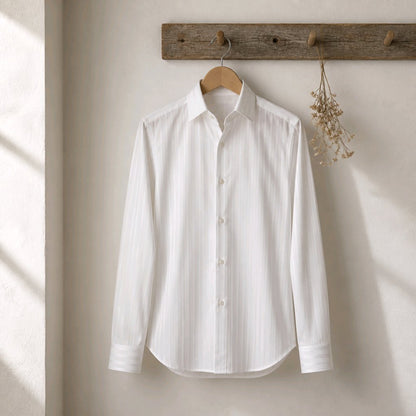 A white long-sleeve button-up shirt with subtle vertical texture, hanging on a wooden hanger from a rustic wooden wall hook against a warm off-white wall.