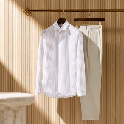 A white long-sleeve button-up shirt with thin vertical stripes hangs on a dark hanger next to a pair of light beige trousers, both hanging from a gold rod against a beige ribbed wall.