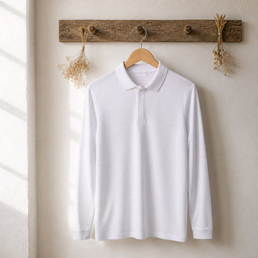 A white long-sleeve polo shirt hanging on a wooden hanger from a rustic wooden wall hook, with dried flowers on either side, against a warm off-white wall.