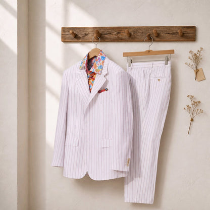 A white pinstripe suit set consisting of a blazer and matching trousers, hanging on wooden hangers from a rustic wooden rack against a textured cream wall.