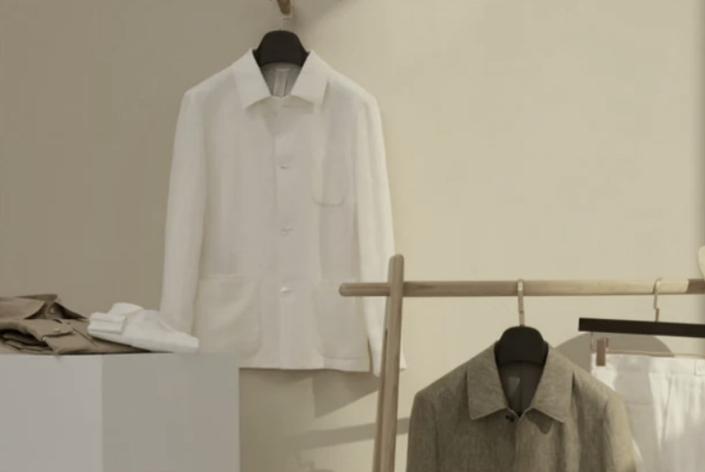 A white, single-breasted, round-necked jacket hanging on a wooden hanger, with a pair of trousers and a waistcoat displayed in the background, all on a light-colored wall.