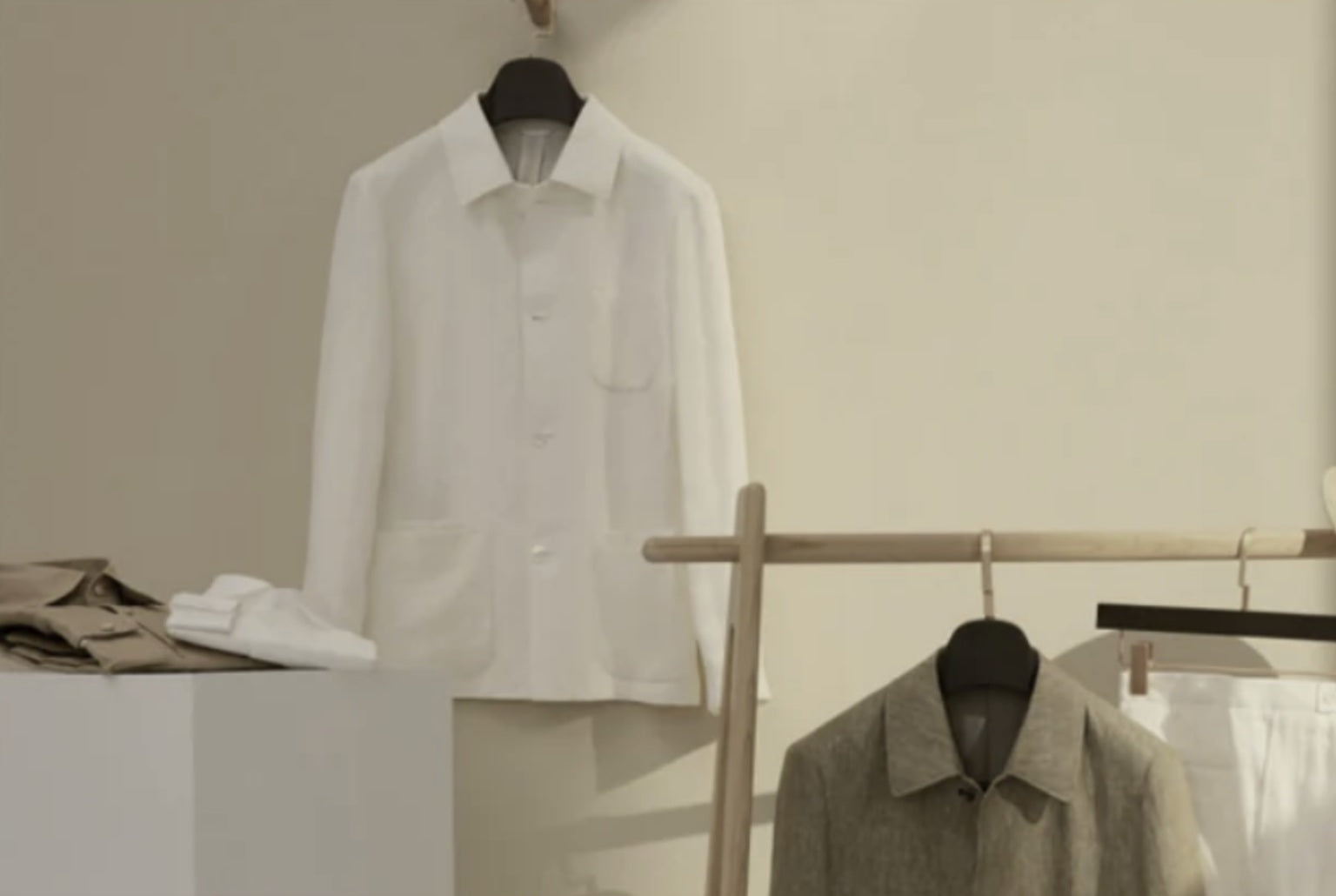 A white, single-breasted, round-necked jacket hanging on a wooden hanger, with a pair of trousers and a waistcoat displayed in the background, all on a light-colored wall.