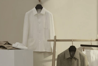 A white, single-breasted, round-necked jacket hanging on a wooden hanger, with a pair of trousers and a waistcoat displayed in the background, all on a light-colored wall.