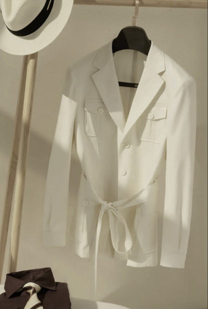 A white, slim-fitted, single-breasted, three-button jacket with a notched lapel and patched flapped breast pocket, displayed on a hanger, with a hat and a waistcoat visible in the background.