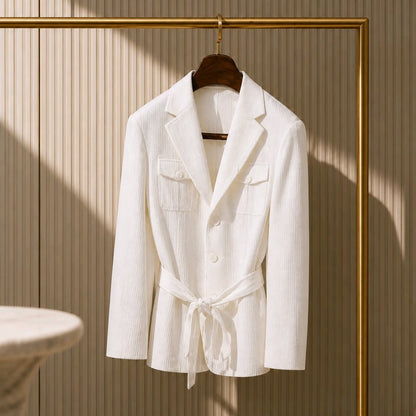 A white textured belted blazer jacket with notch lapels and flap chest pockets, hanging on a wooden hanger from a gold clothing rack against a beige slatted wall.