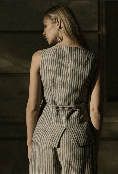 A woman facing away from the camera wears a navy and beige pinstripe blouse with short sleeves and a belt, paired with matching wide-leg trousers. She has brown hair and is accessorized with earrings.